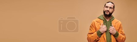 pensive middle eastern man with beard standing in casual attire on beige backdrop, banner