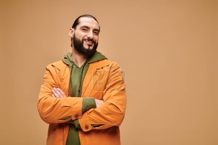 Zufriedener Mann aus dem Mittleren Osten mit Bart steht in lässiger Kleidung mit verschränkten Armen vor beigem Hintergrund