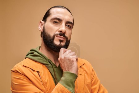 pensive middle eastern man touching beard and standing in casual attire on beige background