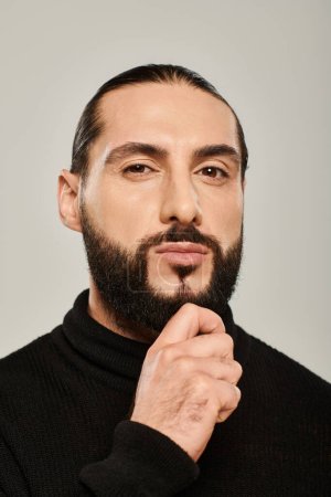 handsome middle eastern man in black turtleneck touching beard on grey background, confidence