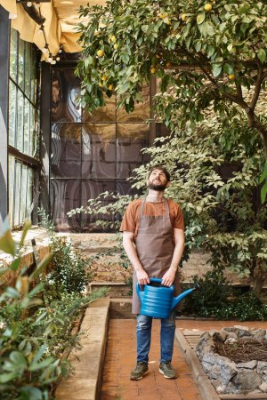 handsome bearded gardener in denim apron holding watering can near trees and plants in greenhouse