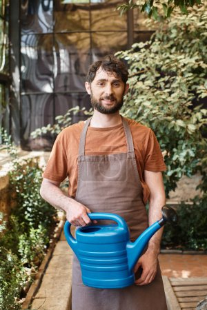 happy bearded gardener in denim apron holding watering can near trees and plants in greenhouse