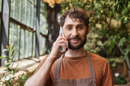 handsome and bearded gardener in apron smiling and talking on smartphone in greenhouse