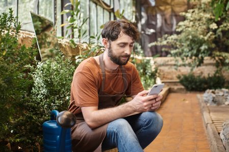 handsome bearded gardener in apron using his on smartphone and sitting around plants in greenhouse