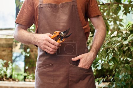 Foto de Cultivado jardinero en delantal de lino con tijeras de jardinería en invernadero, posando con la mano en el bolsillo - Imagen libre de derechos