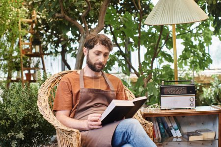 schöner bärtiger Gärtner in Leinenschürze liest Buch und sitzt im Gewächshaus um Pflanzen herum