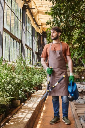 Foto de Jardinero barbudo en guantes y delantal que sostiene grandes tijeras de jardinería alrededor de las plantas en invernadero - Imagen libre de derechos