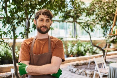 professional headshots, cheerful bearded gardener in apron and gloves posing in greenhouse