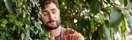 Téléchargez les photos : Beau et barbu jardinier en gants et tablier de lin souriant tout en travaillant en serre, bannière - en image libre de droit