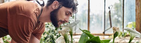 handsome and bearded gardener in linen apron smelling aroma of flower while working in greenhouse