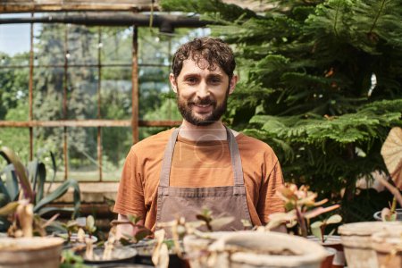 happy bearded gardener in linen apron looking at camera around plants in greenhouse, horticulture