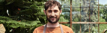happy bearded gardener looking at camera around green plants in greenhouse, horticulture concept