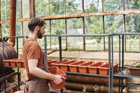 Profi-Gärtner mit Bart in Leinenschürze stellt neue Blumentöpfe ins Gewächshaus