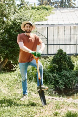 cheerful farmer with beard wearing sun hat and standing with shovel near plants and greenhouse