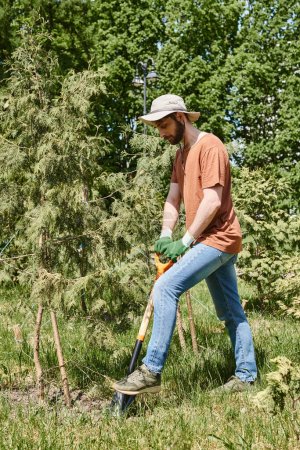 cheerful farmer with beard wearing sun hat and digging with shovel near plants and greenhouse