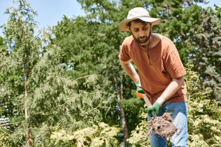 good looking farmer with beard wearing sun hat and digging ground with shovel, plant care