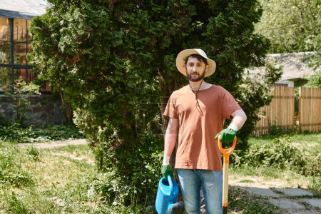 Téléchargez les photos : Beau et barbu jardinier dans chapeau de soleil tenant arrosoir et pelle dans la campagne - en image libre de droit