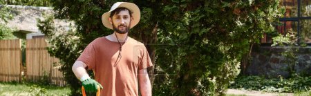 handsome and bearded gardener in sun hat looking at camera and  standing in countryside, banner