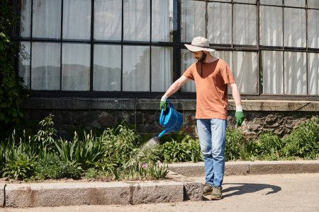 handsome and bearded gardener in sun hat holding watering can and shovel in countryside