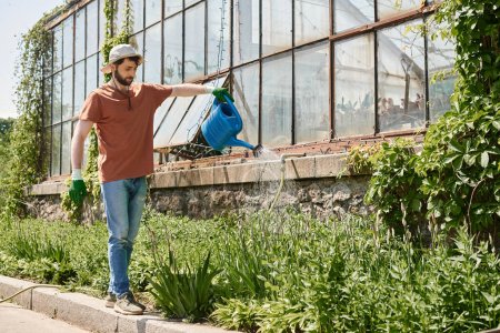 handsome and bearded gardener in sun hat watering plants near greenhouse in countryside