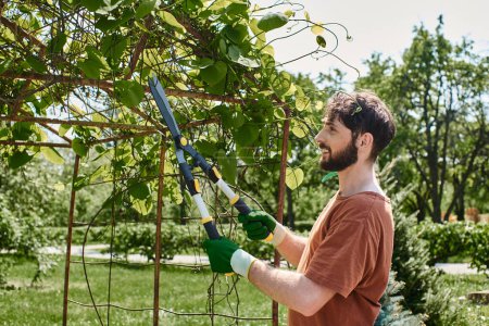 happy bearded gardener in gloves cutting twigs on green tree with big gardening scissors
