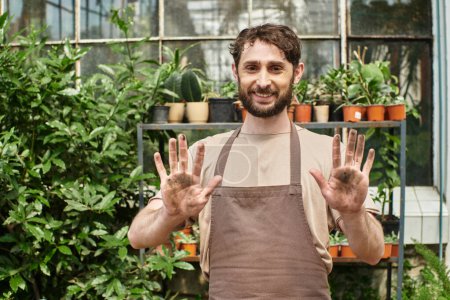 happy bearded gardener in apron showing his dirty hands after working with plants and soil