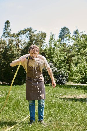 bearded gardener in linen apron drinking water from hose after working in garden, candid photo