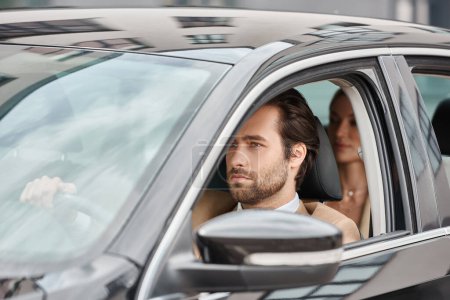 Selbstbewusster bärtiger Mann fährt Luxusauto mit Geschäftsfrau auf Rücksitz vor verschwommenem Hintergrund
