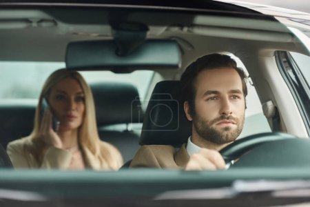 hombre barbudo concentrado conduciendo un coche de lujo con una mujer de negocios rubia hablando por teléfono móvil