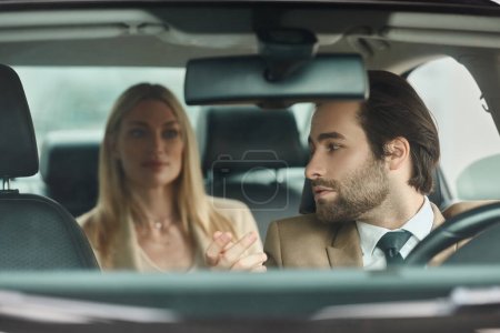 bel homme barbu conduisant une voiture de luxe et parlant à une femme blonde sur le siège arrière, voyage d'affaires