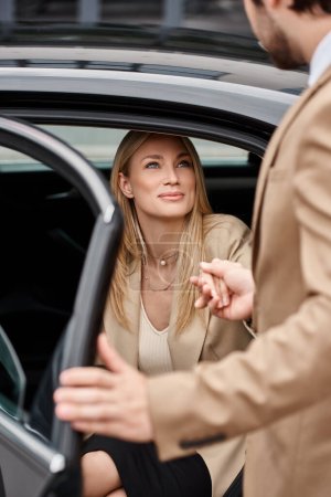 verschwommener Mann hält Hand einer lächelnden blonden Geschäftsfrau in formeller Kleidung beim Aussteigen aus Luxusauto