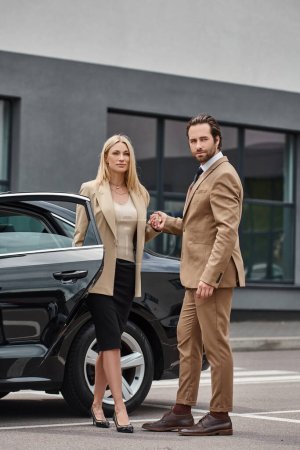 elegant business couple in formal wear holding hands and looking away near car on urban street