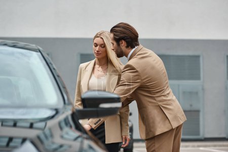 hombre barbudo elegante que abre la puerta trasera del coche de lujo cerca de la elegante mujer de negocios rubia en la calle
