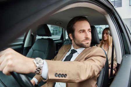 hombre guapo en desgaste formal coche de conducción con atractiva mujer de negocios rubia en el asiento trasero