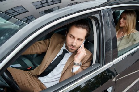 elegant man in formal wear driving car and talking on smartphone near  businesswoman on rear seat