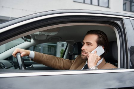 concentrated businessman in beige suit driving car and talking on urban street, entrepreneurship