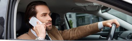 serious businessman in beige suit driving car and talking on urban street, horizontal banner