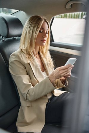 élégante femme d'affaires blonde dans la messagerie formelle d'usure sur le téléphone mobile dans la voiture de luxe sur la rue de la ville