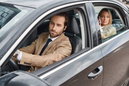 handsome and elegant man driving car near attractive blonde businesswoman talking on smartphone