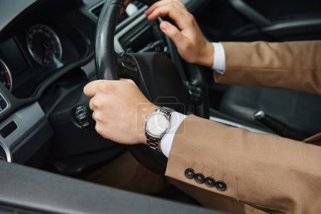 Ausgeschnittene Ansicht eines erfolgreichen Geschäftsmannes in beigem Anzug und Armbanduhr, der Luxuswagen auf der Straße fährt