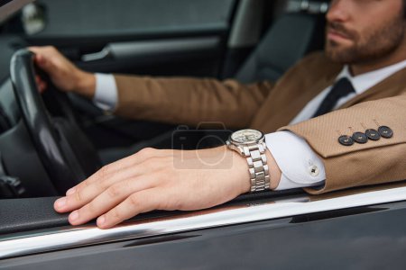 Ausgeschnittene Ansicht eines selbstbewussten Geschäftsmannes in beigem Anzug und Armbanduhr, der Luxuswagen auf der Straße fährt