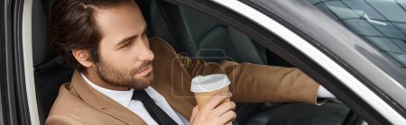 bearded businessman in beige elegant suit holding coffee to go and driving car on street,  banner