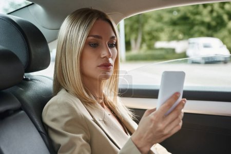 Blonde Geschäftsfrau in eleganter formaler Kleidung surft im Internet auf dem Handy, während sie im Auto sitzt