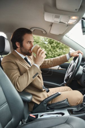 stylish bearded businessman in beige suit drinking coffee to go and driving car on urban street