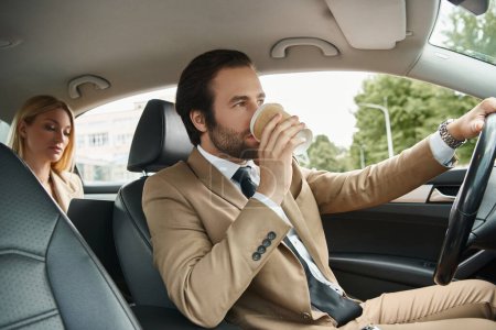 stylish man in formal wear drinking coffee to and driving car with blonde businesswoman on rear seat