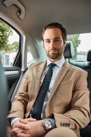 ruhiger und selbstbewusster Geschäftsmann im beige-eleganten Anzug, der in einem Luxusauto auf der Straße in die Kamera blickt