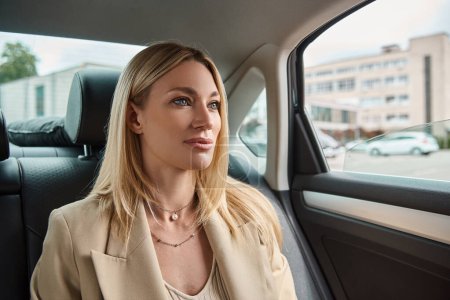 attractive blonde businesswoman in beige elegant blazer traveling in luxury car on city street