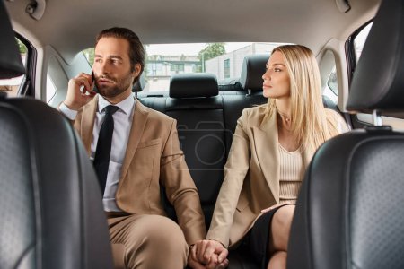 stylish blonde businesswoman holding hands with elegant man talking on smartphone in car, attraction