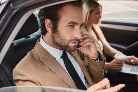 Mann in offizieller Kleidung unterhält sich mit Smartphone in der Nähe von Geschäftsfrau mit Coffee to go und Laptop im Auto
