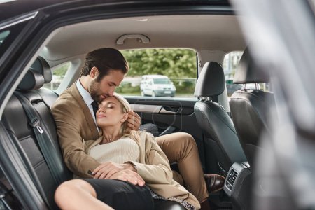stylish businessman with closed eyes embracing sensual blonde woman in luxury car, love affair